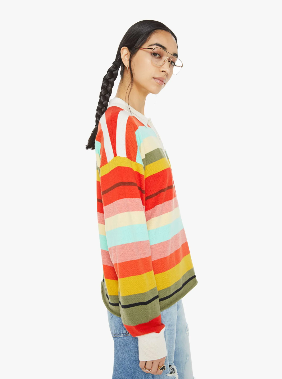 A person with glasses stands against a plain background, wearing The Penalty Kick Rainbow sweater by Mother, featuring vibrant rainbow stripes. They pair it with light blue jeans, and their braided hair complements the relaxed expression reminiscent of an athletic-inspired polo style as they look to the side.