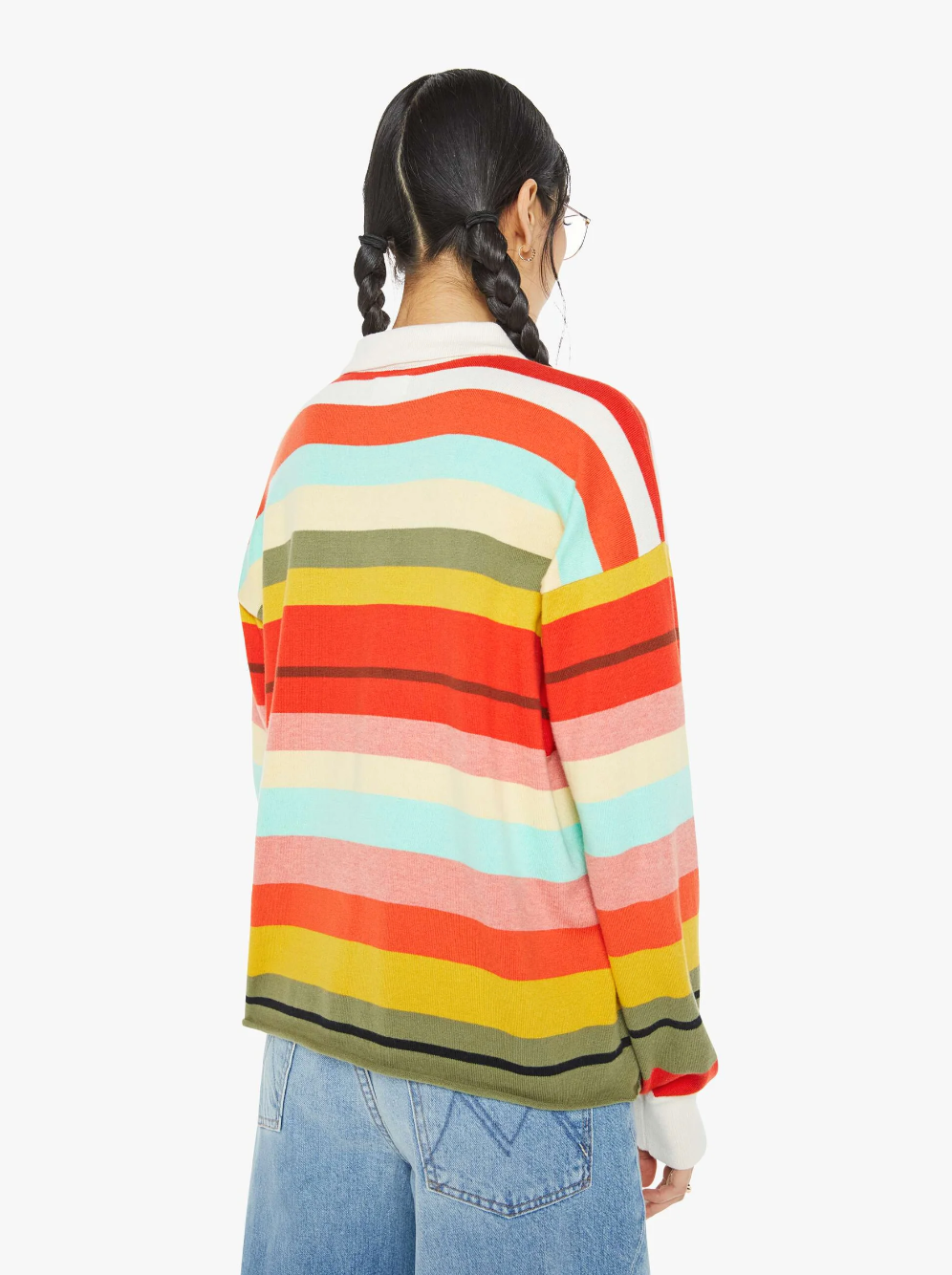 A person with braided hair stands with their back to the camera, wearing Mother’s "The Penalty Kick Rainbow," an oversized fit sweater featuring vibrant rainbow stripes of red, orange, blue, and green. Paired with light blue jeans against a plain white background, the colorful ensemble creates a striking visual.