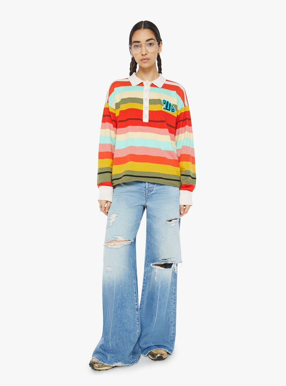 A person stands against a plain background wearing "The Penalty Kick Rainbow" sweater by Mother, featuring vibrant rainbow stripes and paired with light blue ripped jeans. The look is completed with large round glasses and long brown hair tied back.