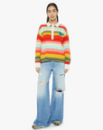 A person stands against a plain background wearing "The Penalty Kick Rainbow" sweater by Mother, featuring vibrant rainbow stripes and paired with light blue ripped jeans. The look is completed with large round glasses and long brown hair tied back.