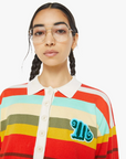 A person with braided hair and glasses wears "The Penalty Kick Rainbow" polo shirt by Mother, featuring vibrant stripes in red, orange, yellow, green, and blue. The shirt’s colorful design is enhanced by a white collar and a blue embroidered letter on the chest.