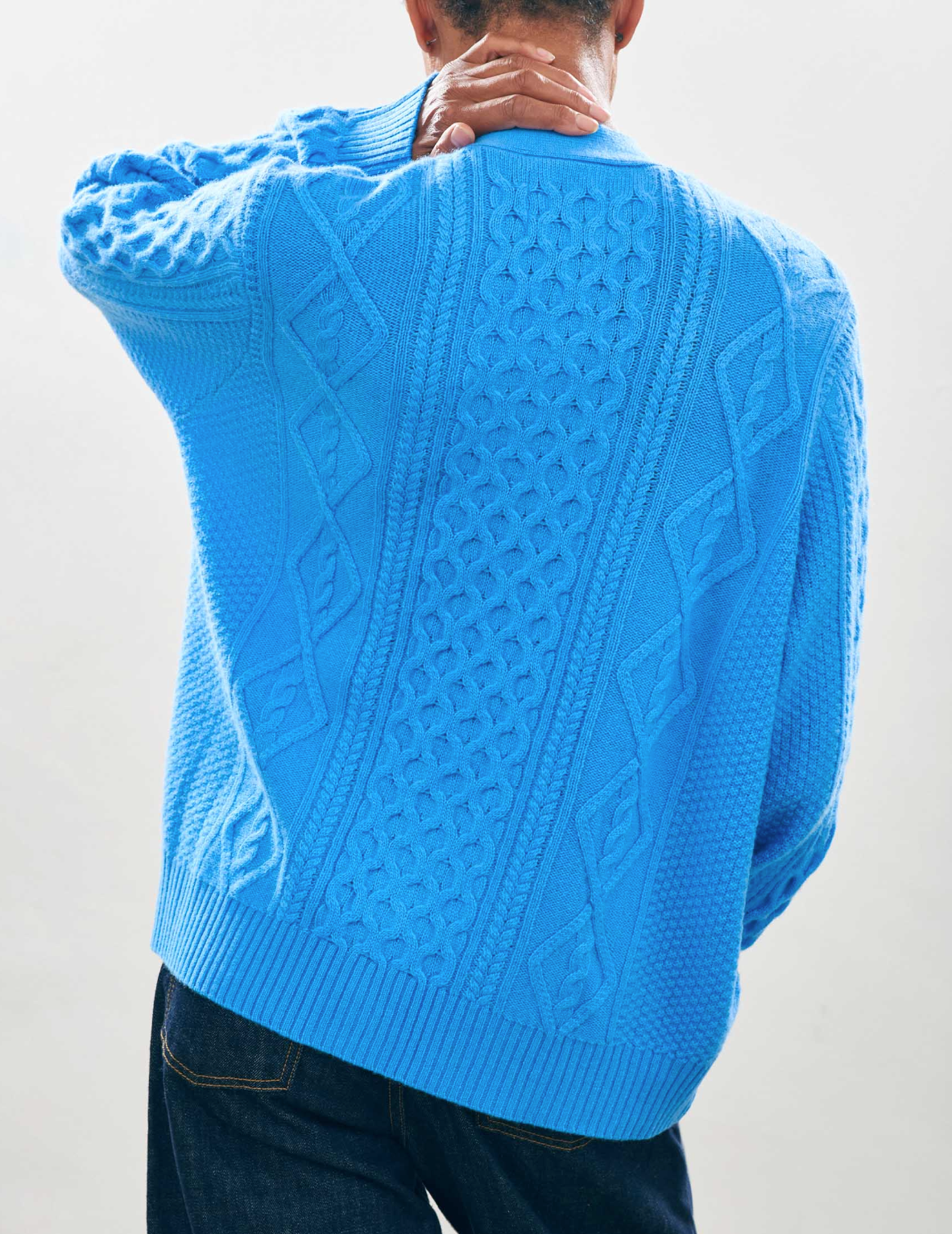 A person wearing a bright blue, textured knit Aran Cardigan from Jumper 1 2 3 4/CR2, showcasing intricate patterns and an oversized fit, stands facing away with one hand on the back of their neck against a plain background.