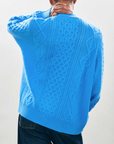 A person wearing a bright blue, textured knit Aran Cardigan from Jumper 1 2 3 4/CR2, showcasing intricate patterns and an oversized fit, stands facing away with one hand on the back of their neck against a plain background.