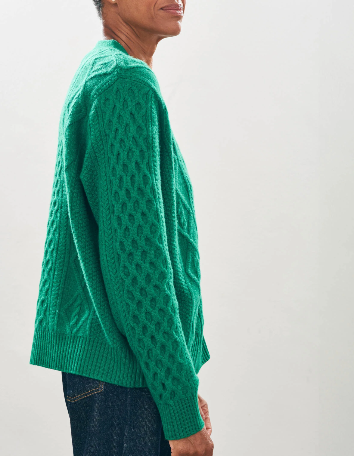 Against a plain background, someone stands facing away from the camera, showcasing a vibrant green Aran Cardigan with a visible right arm. This oversized sweater by Jumper 1 2 3 4/CR2 has a classic vee neck design and features an intricate, textured diamond-shaped pattern on the back.