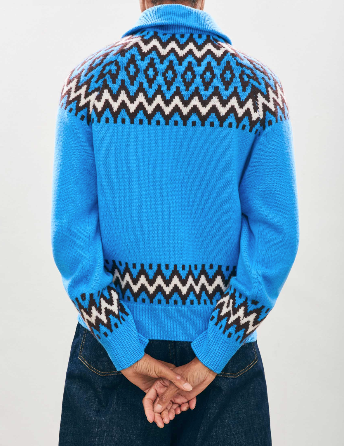 A person wearing an oversize fit Ski Zipup in bright blue, featuring black and white geometric patterns, stands with their back to the camera, hands clasped behind their back. This product from Jumper 1 2 3 4/CR2 has a high collar that evokes the style of cashmere wool cardigans, paired with dark pants.