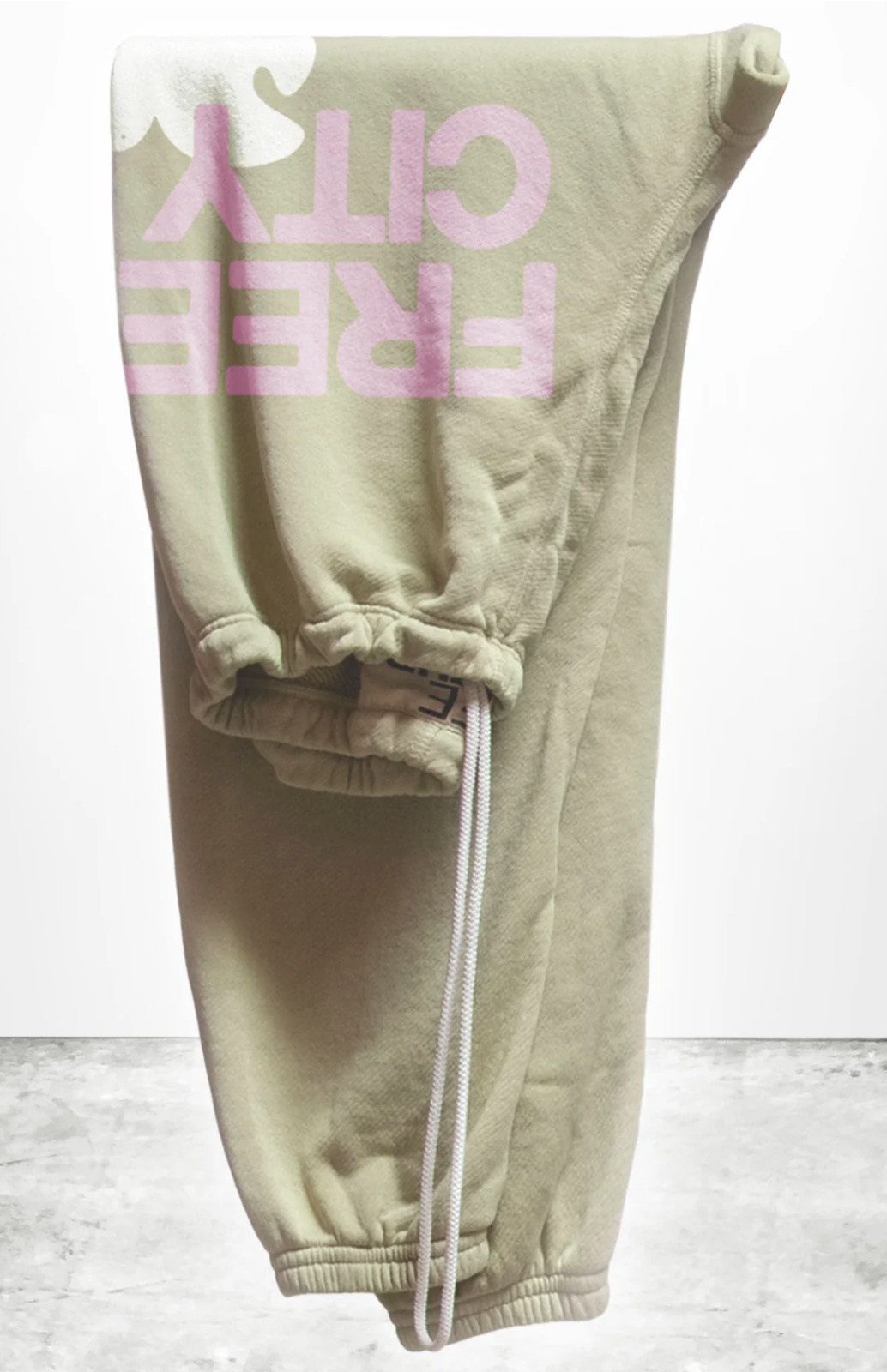 The SUPERFLUFF Lux OG Sweatpants from Free City (Sparrow, LLC) showcase pink and white hand-screen printed "FREE CITY" text on the side against a plain background. These olive green unisex sweatpants are made from lightweight French terry and feature an elastic waistband with a drawstring, as well as gathered cuffs for added comfort.