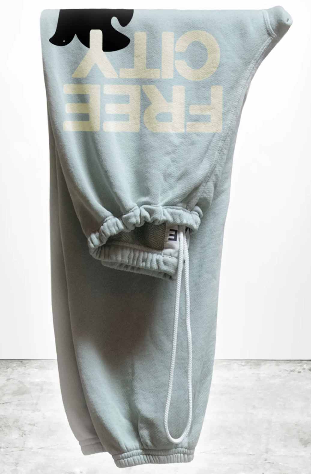 Displayed is a folded pair of light gray, unisex SUPERFLUFF Lux OG Sweatpants by Free City (sparrow, LLC), made from lightweight French terry. The drawstring waist highlights the partially visible "FREE CITY" design in bold yellowish letters. These stylish essentials are set against a minimalistic concrete floor backdrop.