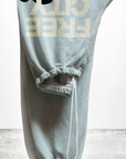 Displayed is a folded pair of light gray, unisex SUPERFLUFF Lux OG Sweatpants by Free City (sparrow, LLC), made from lightweight French terry. The drawstring waist highlights the partially visible "FREE CITY" design in bold yellowish letters. These stylish essentials are set against a minimalistic concrete floor backdrop.