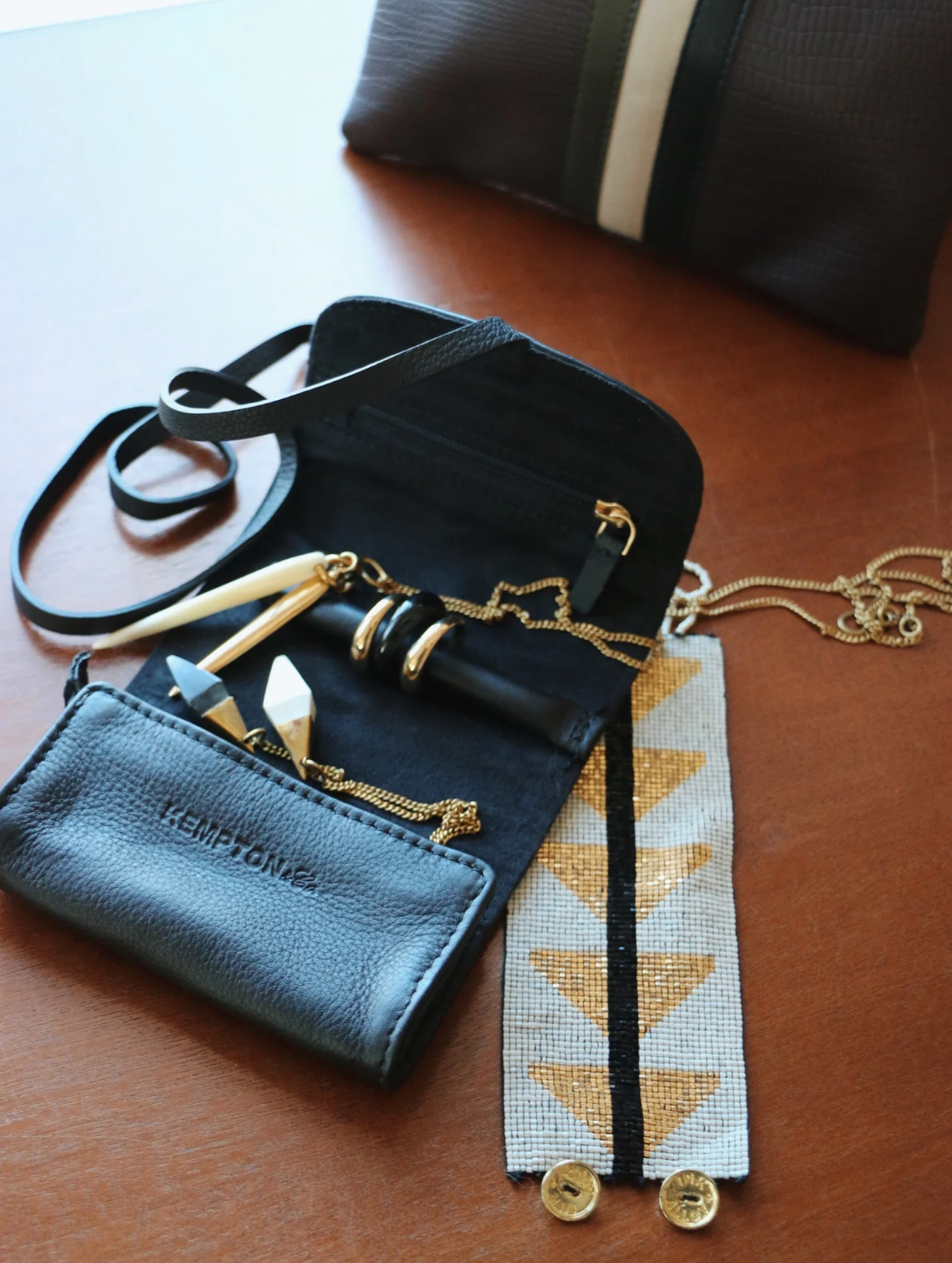 On a wooden surface, a black cowhide handbag holds gold pens, along with a segmented black and gold cosmetic item and a piece of embroidered fabric. Nearby, the Kempton & Co. Black Leather Jewelry Roll, perfect for traveling, lies alongside a small black pouch with gold text and a brown striped pouch.