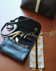 On a wooden surface, a black cowhide handbag holds gold pens, along with a segmented black and gold cosmetic item and a piece of embroidered fabric. Nearby, the Kempton & Co. Black Leather Jewelry Roll, perfect for traveling, lies alongside a small black pouch with gold text and a brown striped pouch.