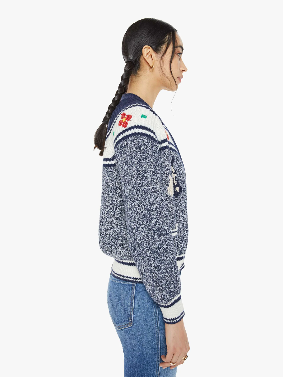 A woman in profile features braided hair, wearing "The Try Outs" crewneck cardigan sweater by Mother, showcasing a marled navy and white design with vibrant floral accents. The cardigan is crafted from a cotton and alpaca blend, complete with striped cuffs and hem, and she pairs it elegantly with jeans against a plain white background.