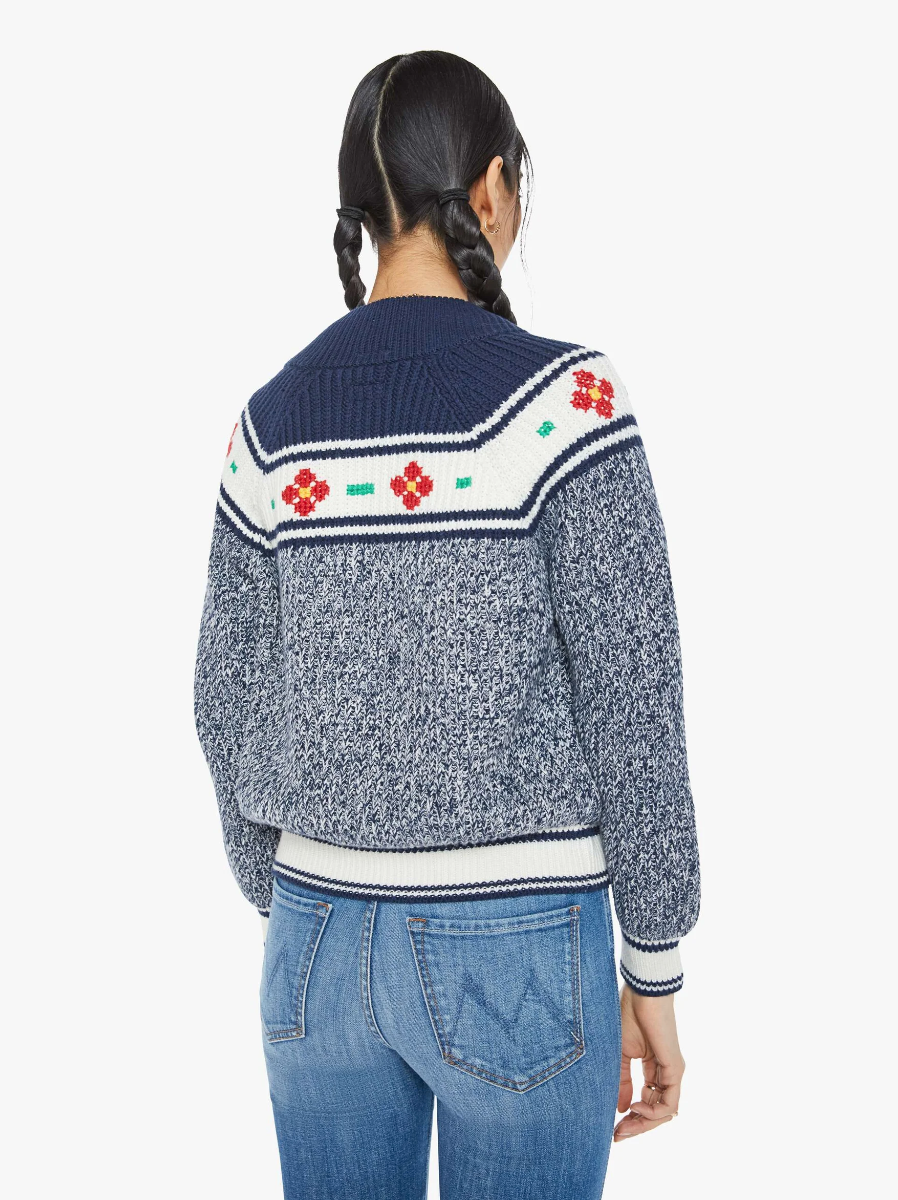 Rear view of a person with braided hair, wearing "The Try Outs" cardigan sweater by Mother in marled navy cotton and alpaca, featuring floral patterns on the shoulders. They also have on blue jeans. The background is plain white.