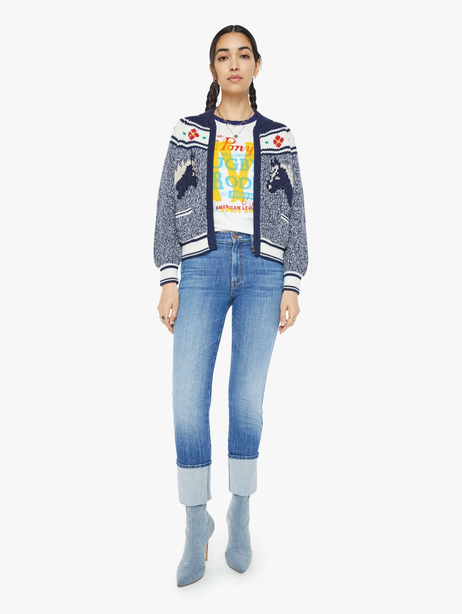 A woman stands against a plain white background, wearing "The Try Outs" jacket by Mother over a colorful graphic T-shirt, paired with marled navy blue cuffed jeans and gray ankle boots.