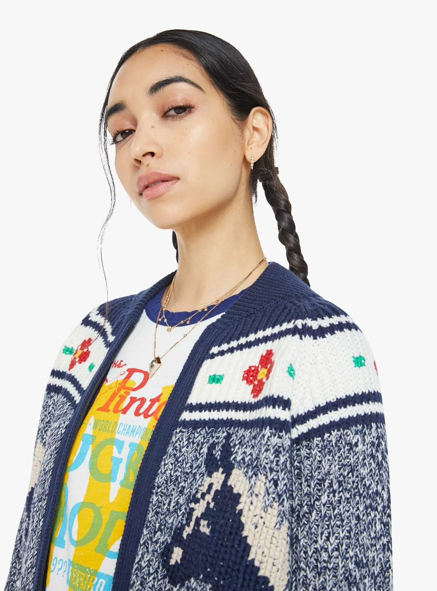 A person with a braided hairstyle is wearing a colorful graphic t-shirt and "The Try Outs" marled navy and white crewneck cardigan sweater from Mother, which is crafted from a cotton and alpaca blend. The cardigan features intricate blue, white, and red patterns set against a plain white background.