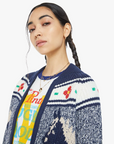 A person with a braided hairstyle is wearing a colorful graphic t-shirt and "The Try Outs" marled navy and white crewneck cardigan sweater from Mother, which is crafted from a cotton and alpaca blend. The cardigan features intricate blue, white, and red patterns set against a plain white background.