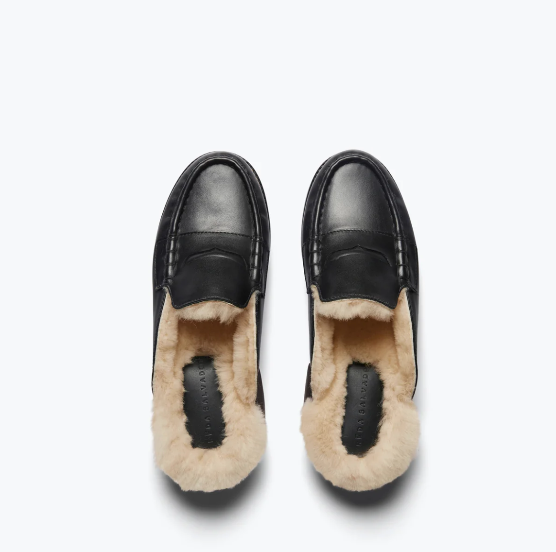 From an overhead perspective, the TEMI SHEARLING PENNY LOAFER MULE by Freda Salvador showcases a sleek, polished exterior made from Italian leather in black and a cozy interior lined with tan ethically sourced shearling.