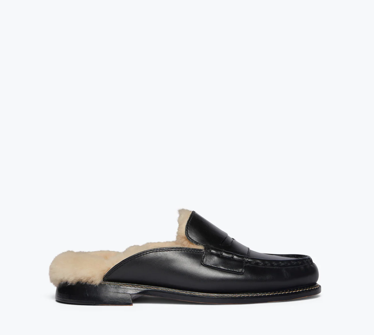 The TEMI SHEARLING PENNY LOAFER MULE by Freda Salvador is shown from the side on a plain white background, featuring a classic loafer design made from Italian leather with stitching details. This black leather slip-on shoe reveals cream-colored, ethically sourced shearling lining.