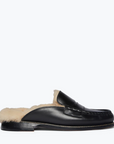 The TEMI SHEARLING PENNY LOAFER MULE by Freda Salvador is shown from the side on a plain white background, featuring a classic loafer design made from Italian leather with stitching details. This black leather slip-on shoe reveals cream-colored, ethically sourced shearling lining.