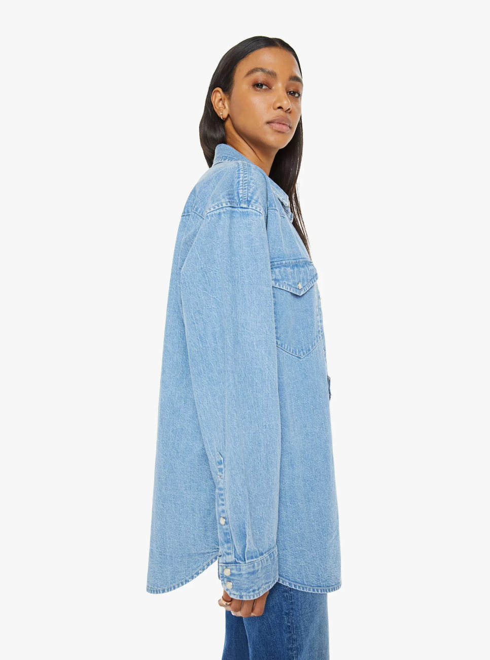 A person with long dark hair wears "The Exes And The Ohs," an oversized denim button-up from Mother, featuring a vintage-blue wash and faux pearl snaps, standing sideways against a plain white background.