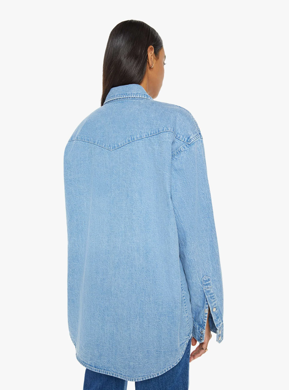 A person wearing "The Exes And The Ohs" denim jacket by Mother in vintage-blue wash with faux pearl snaps is facing away, showcasing the back. The jacket includes long sleeves and a classic collar. Their hair is down, and they are paired with blue jeans against a plain white background.