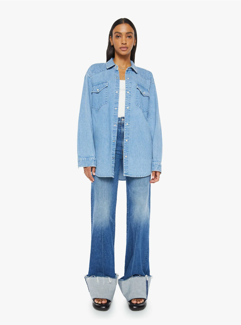 A person wearing "The Exes And The Ohs" by Mother, an oversized denim button-up paired with wide-legged jeans featuring cuffs, vintage-blue wash, and faux pearl snaps, poses against a plain white background. The ensemble exudes a stylish and casual vibe.