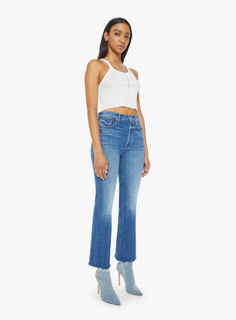 A person wearing a white sleeveless top, high-rise The Tripper Ankle Fray jeans by Mother, and light blue heeled boots stands against a plain white background. These premium jeans, crafted in Los Angeles, feature a slight flare at the bottom.