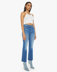 A person wearing a white sleeveless top, high-rise The Tripper Ankle Fray jeans by Mother, and light blue heeled boots stands against a plain white background. These premium jeans, crafted in Los Angeles, feature a slight flare at the bottom.