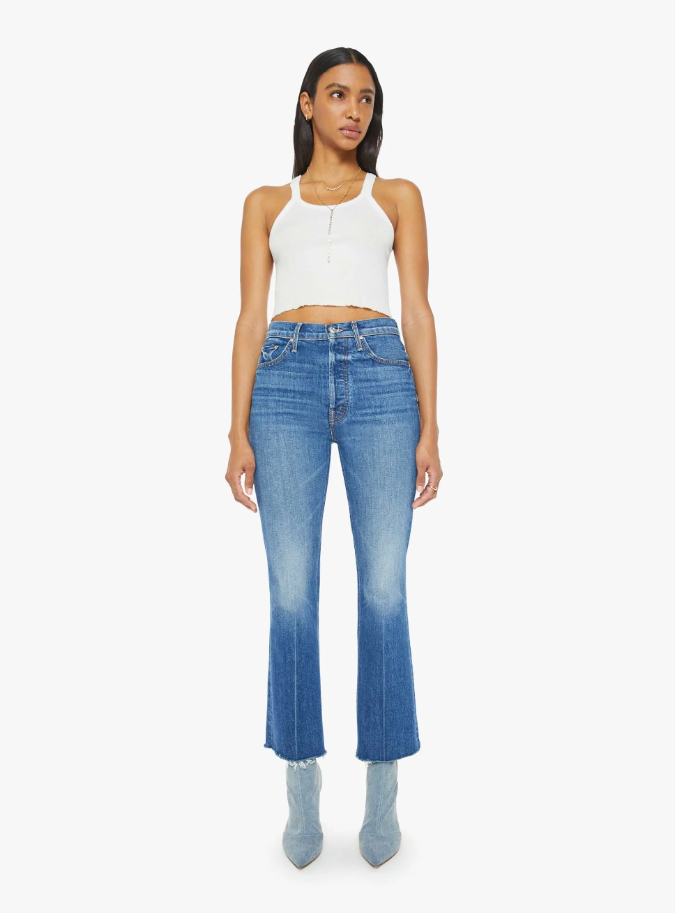 A person stands against a white background wearing a white tank top, The Tripper Ankle Fray high-rise flare jeans by Mother in a mid-blue wash, and blue ankle boots. They have long dark hair and are gazing forward with a neutral expression.