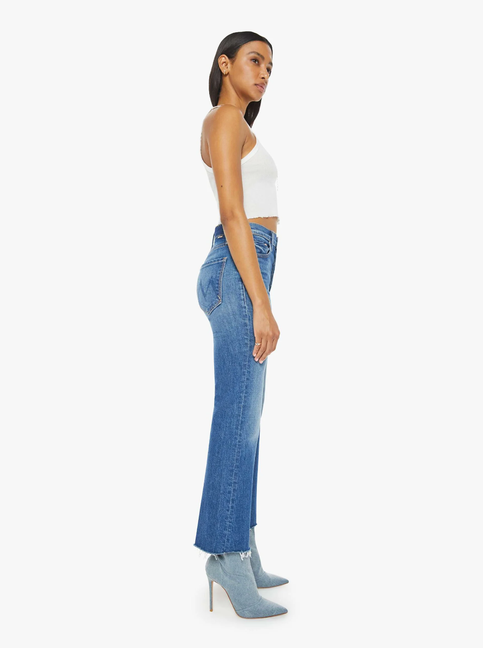 A person with long dark hair is standing in profile, wearing a white sleeveless top paired with The Tripper Ankle Fray jeans by Mother. These high-rise flare jeans come in a mid-blue wash and are made from semi-rigid denim. The outfit is complemented by light blue high-heeled ankle boots, set against a plain white background.