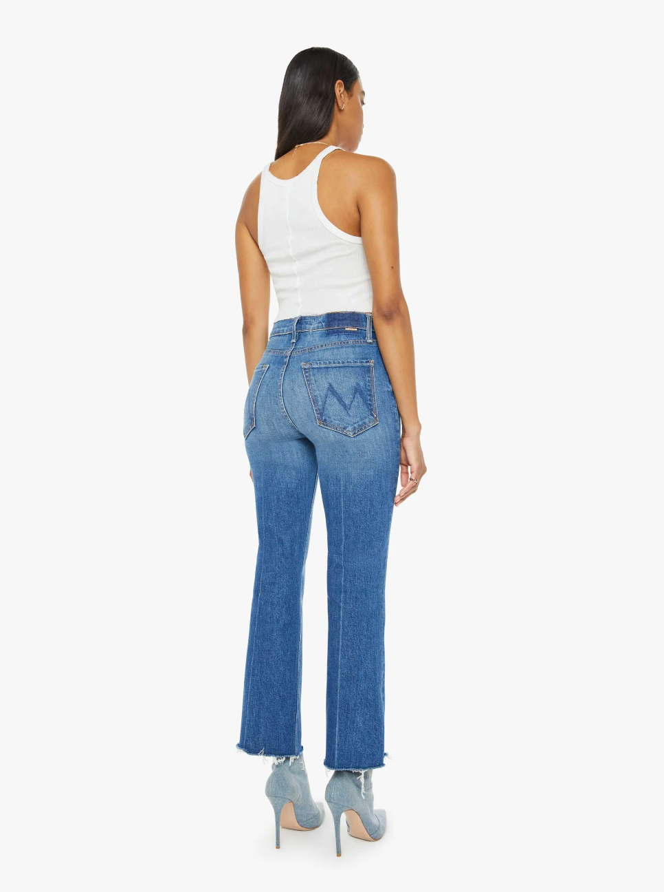 A person with long hair stands facing away, wearing a white tank top, The Tripper Ankle Fray high-rise flare jeans by Mother in SUPERIOR denim with frayed hems, and light blue heels. The mid-blue wash of the jeans complements the minimalist aesthetic against the plain white background.