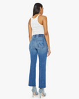 A person with long hair stands facing away, wearing a white tank top, The Tripper Ankle Fray high-rise flare jeans by Mother in SUPERIOR denim with frayed hems, and light blue heels. The mid-blue wash of the jeans complements the minimalist aesthetic against the plain white background.