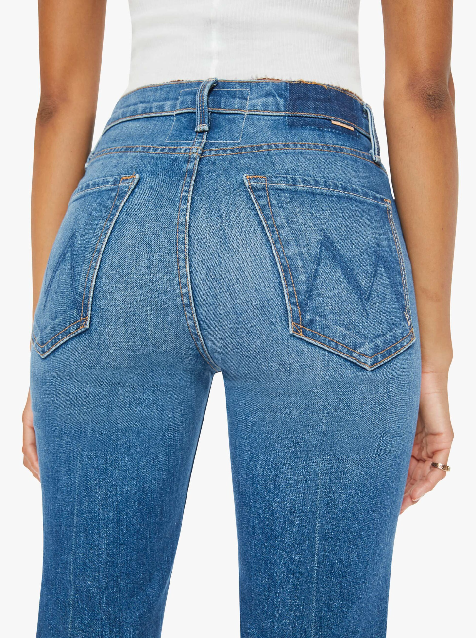 A person is shown from the rear wearing The Tripper Ankle Fray high-rise flare jeans by Mother, crafted in Los Angeles. These jeans feature back pockets with an embroidered design and are paired with a white top and a small ring on the person's left hand.