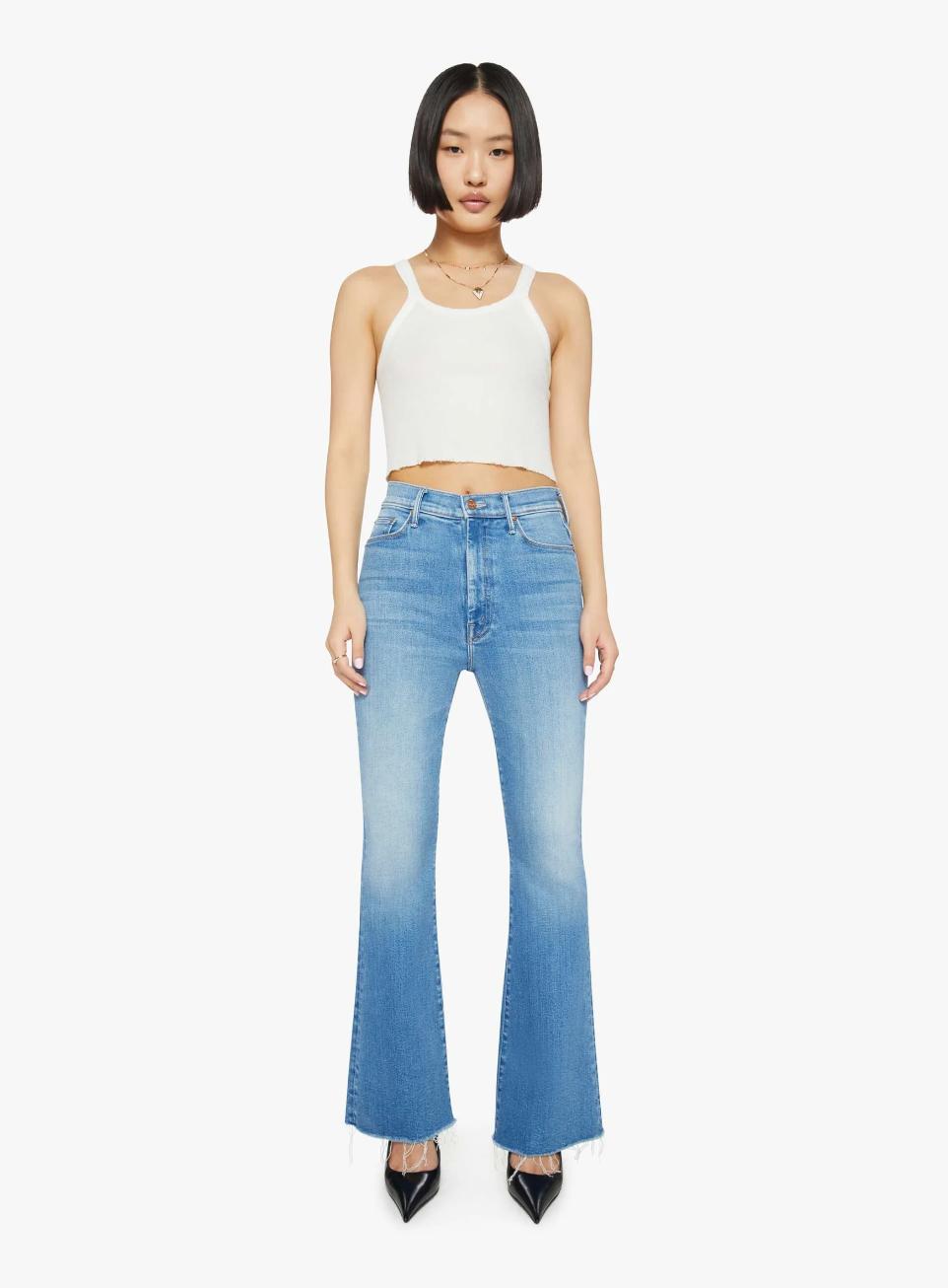 A person with short black hair wears Mother’s Lil HW Weekender Fray, a pair of flared blue jeans made of stretch denim, paired with a white tank top and black pointed shoes. They stand against a plain white background.