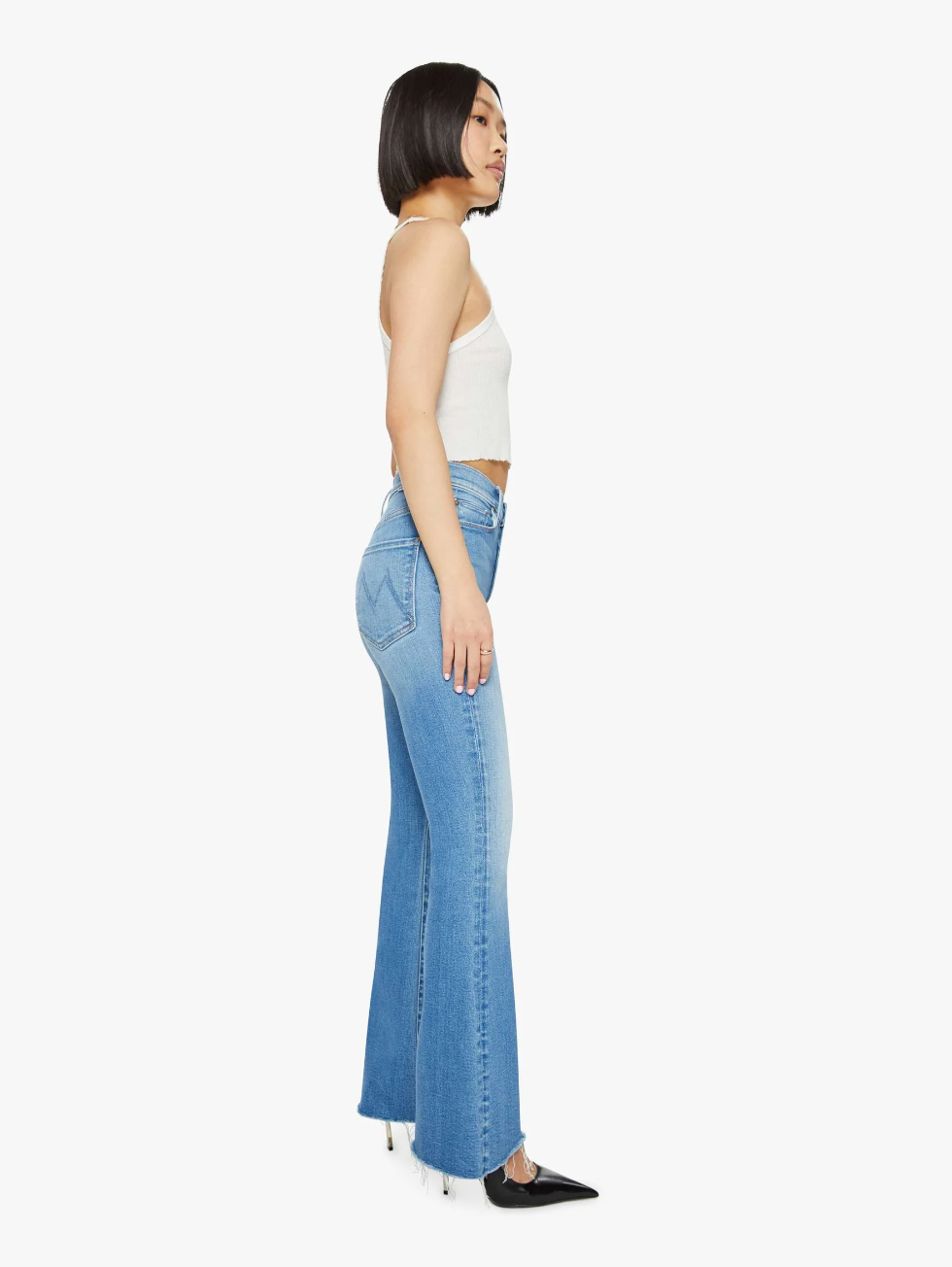 A person with a bob haircut is wearing a white sleeveless top and the Lil HW Weekender Fray flare jeans by Mother in a medium blue wash, paired with black pointed shoes, standing in profile against a white background.