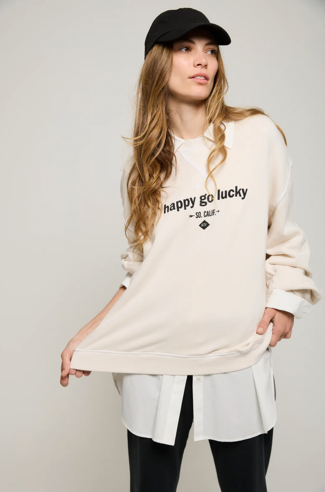 A woman with long hair, wearing a black cap, is dressed in an oversized "Happy Go Lucky BF Sweatshirt" from ATF Clothing, made of brushed terry. The beige sweatshirt features the "Happy Go Lucky" graphic and is layered over a white shirt. She pairs it with black pants as she poses against a plain background, slightly pulling on the hem.