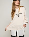 A woman with long hair, wearing a black cap, is dressed in an oversized "Happy Go Lucky BF Sweatshirt" from ATF Clothing, made of brushed terry. The beige sweatshirt features the "Happy Go Lucky" graphic and is layered over a white shirt. She pairs it with black pants as she poses against a plain background, slightly pulling on the hem.