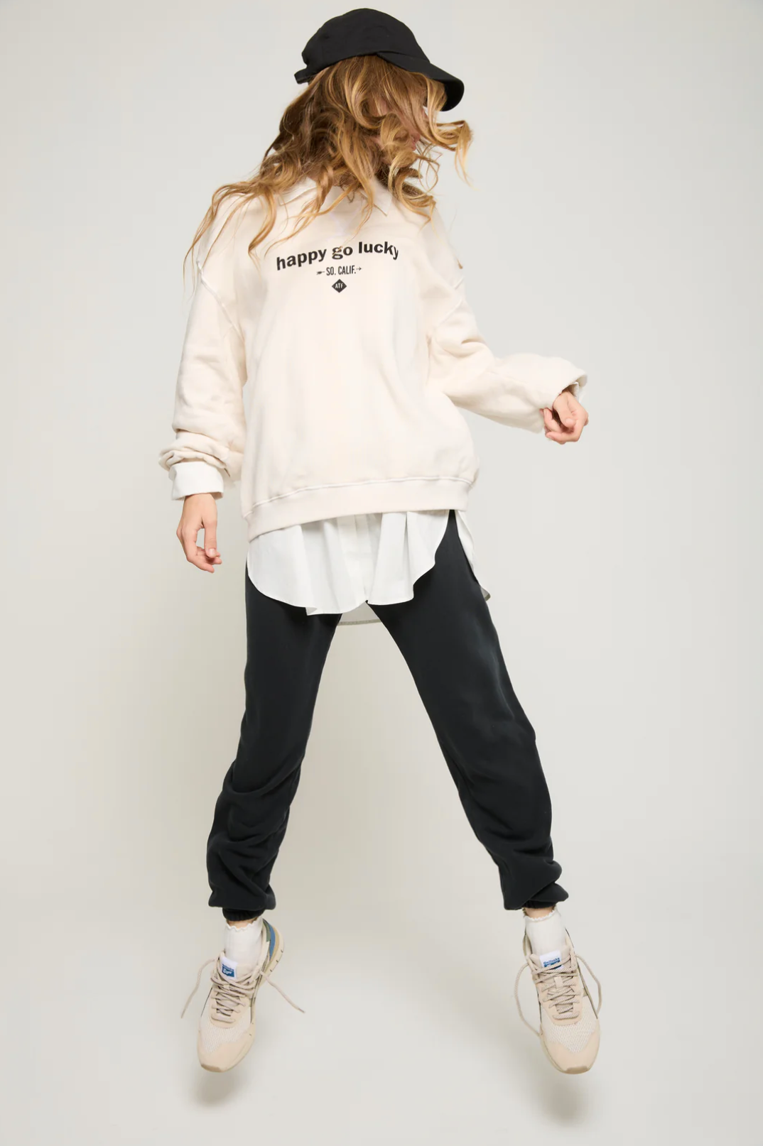 A person sporting the "Happy Go Lucky BF Sweatshirt" by ATF Clothing—an oversized pullover in black featuring the signature graphic—with black pants and white sneakers, is captured mid-jump against a plain background. The beige brushed terry fabric enhances comfort, with a white shirt subtly showing beneath the sweatshirt.