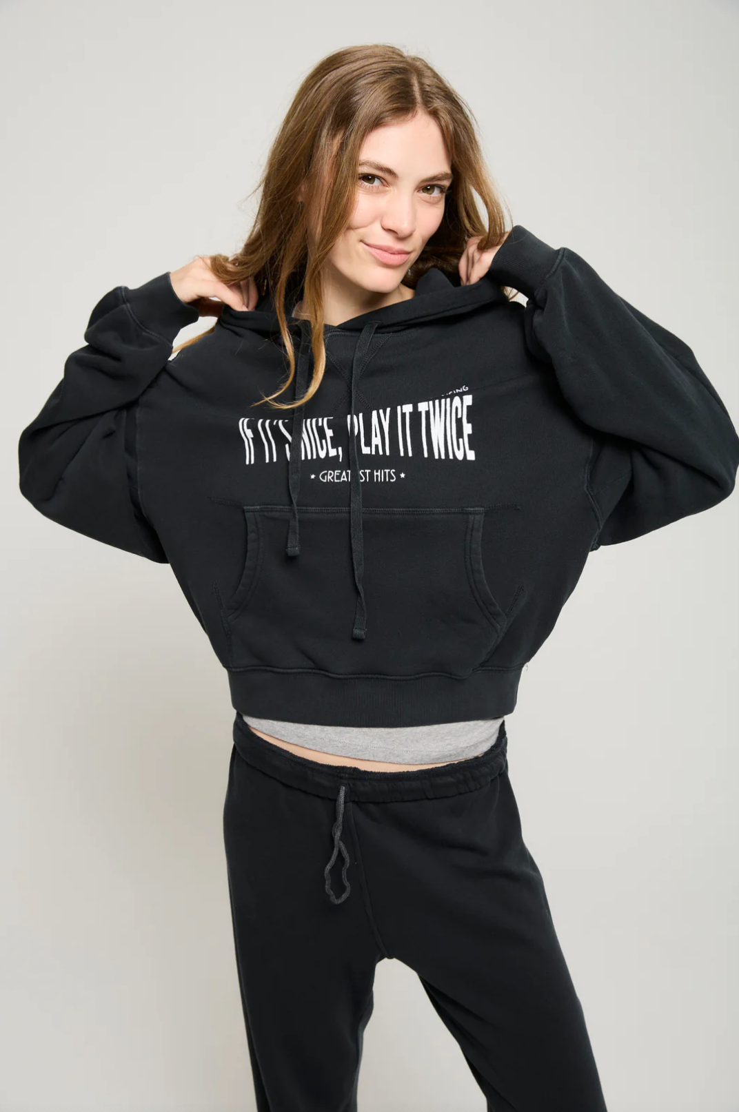 A person with long hair wears an ultra-soft black cropped hoodie from ATF Clothing, featuring the text "If It's Nice, Play It Twice, Greatest Hits," paired perfectly with black pants. They are smiling and holding the Play it Twice Cropped Hoodie with both hands while standing against a plain light gray background, showcasing MADE IN USA quality through their style.