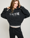 A person with long hair wears an ultra-soft black cropped hoodie from ATF Clothing, featuring the text "If It's Nice, Play It Twice, Greatest Hits," paired perfectly with black pants. They are smiling and holding the Play it Twice Cropped Hoodie with both hands while standing against a plain light gray background, showcasing MADE IN USA quality through their style.