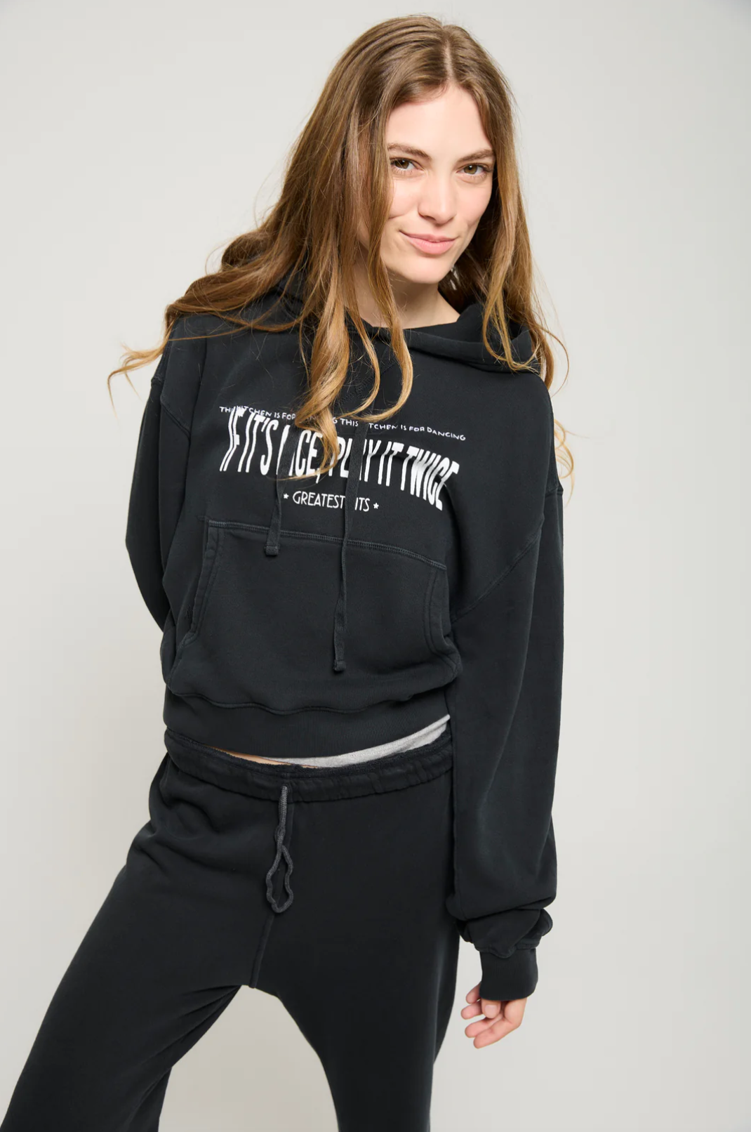 A person with long hair is wearing the Play it Twice Cropped Hoodie by ATF Clothing, which features an embroidered graphic. They pair this black hoodie with black pants and stand against a plain background in a relaxed, casual pose.