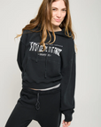 A person with long hair is wearing the Play it Twice Cropped Hoodie by ATF Clothing, which features an embroidered graphic. They pair this black hoodie with black pants and stand against a plain background in a relaxed, casual pose.