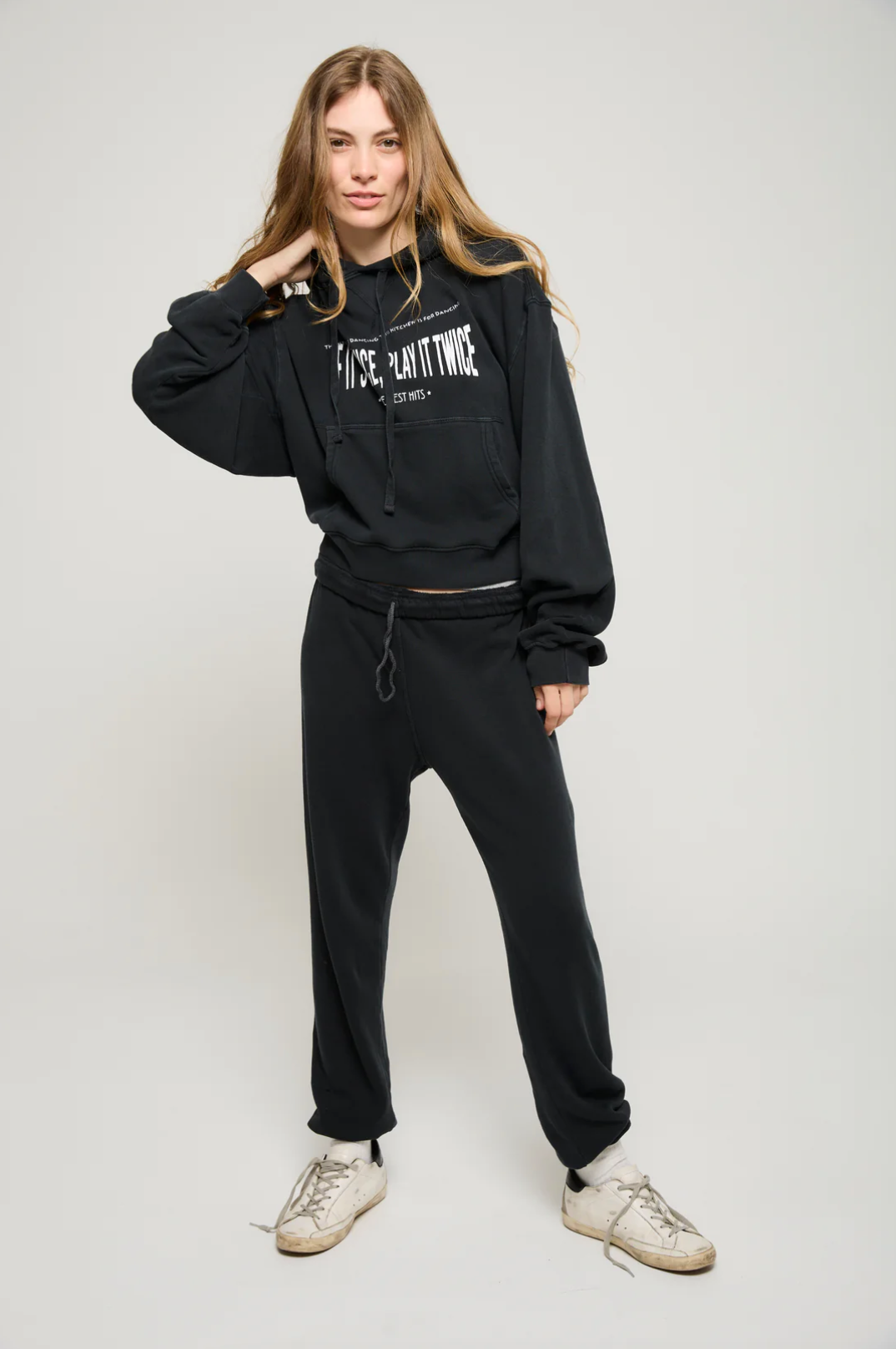 A person is sporting the Play it Twice Cropped Hoodie by ATF Clothing, a black cropped hoodie made from brushed terry fabric with "FUSE PLANT THREE" graphic printed on it, paired with matching black sweatpants. They pose against a plain backdrop, one hand touching their hair, while wearing white sneakers.