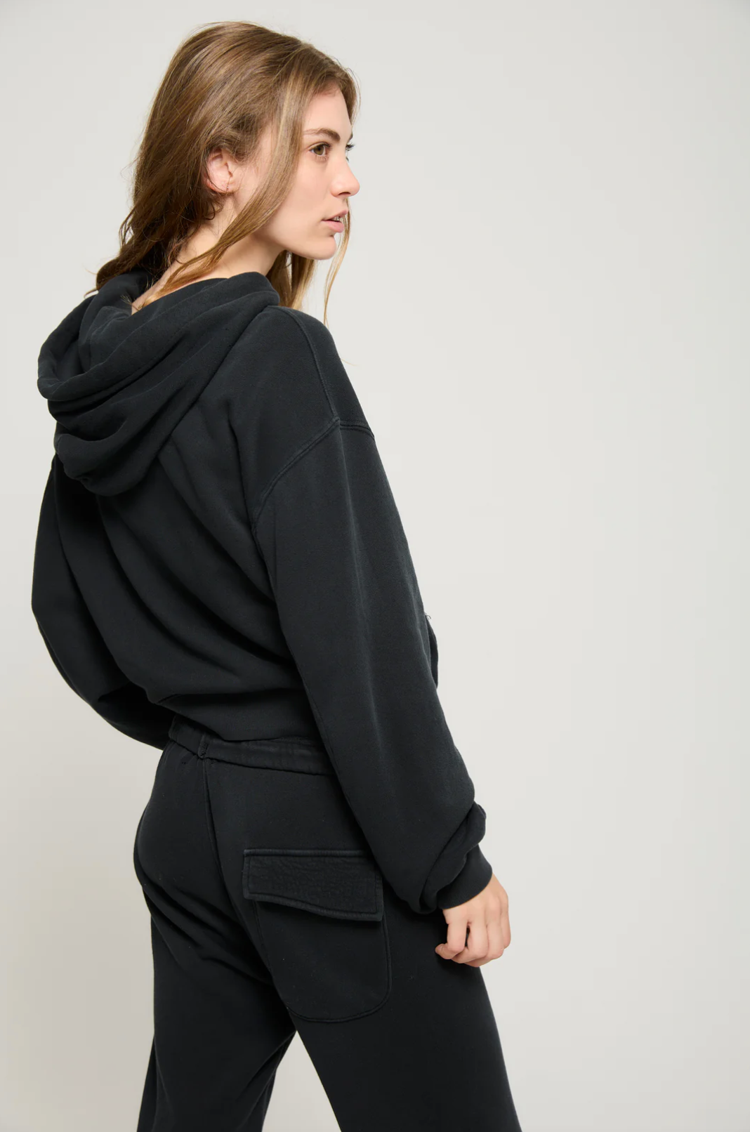 A person with long hair is wearing the Play it Twice Cropped Hoodie by ATF Clothing, made from brushed terry, and black pants. They stand sideways, looking slightly to the left against a plain, light-colored background.