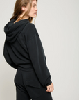 A person with long hair is wearing the Play it Twice Cropped Hoodie by ATF Clothing, made from brushed terry, and black pants. They stand sideways, looking slightly to the left against a plain, light-colored background.