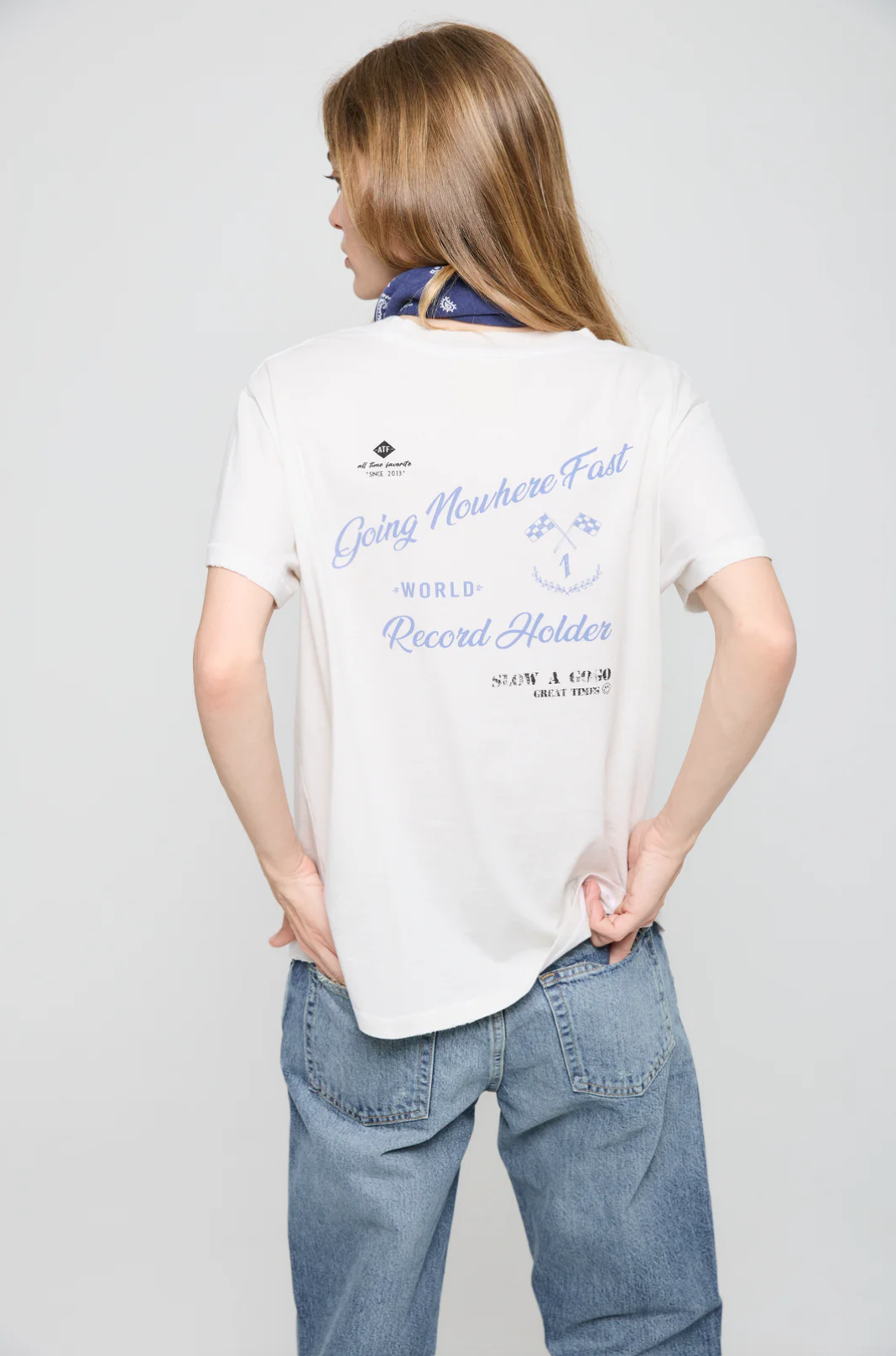 A person with long hair wearing a white Going Nowhere Fast SS Crew T-shirt from ATF Clothing and blue jeans stands facing away. The shirt showcases a "Going Nowhere Fast" graphic adorned with racing flag print details. To complement the casual look, they have a blue bandana tied around their neck.