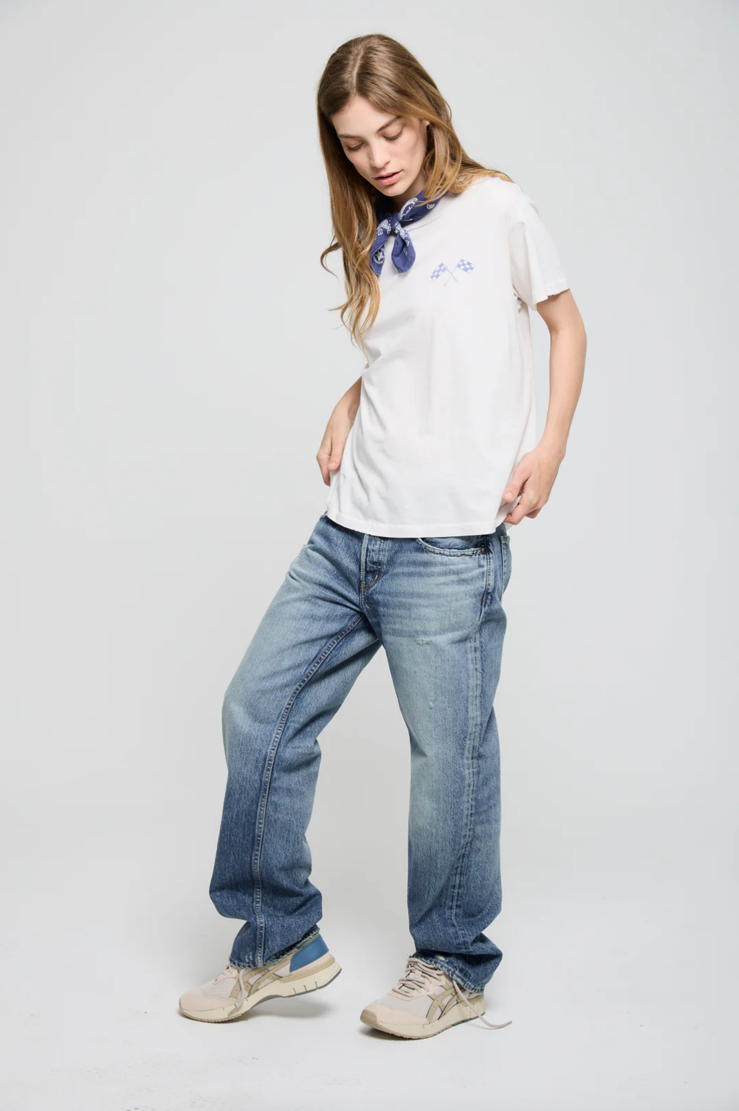 A person with long hair wearing an ATF Clothing "Going Nowhere Fast SS Crew" graphic tee, blue jeans, and beige sneakers stands against a plain background. They have a blue bandana around their neck and are looking down with one hand in a pocket.