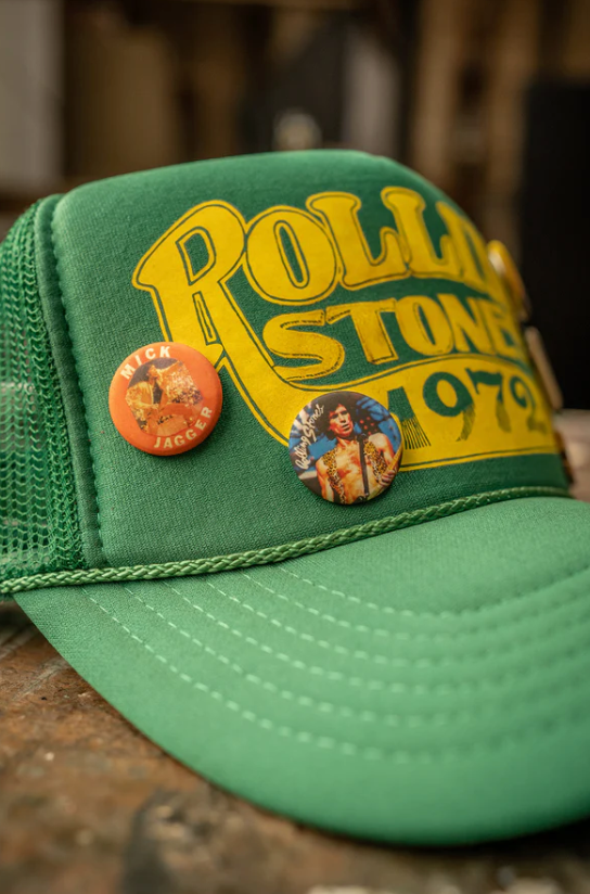 The Made Worn Rolling Stones 1972 Hat is a green trucker hat with yellow text and an adjustable back strap for a perfect fit. It's adorned with two custom pins: one of Mick Jagger and another featuring a person in colorful attire. One size fits most.