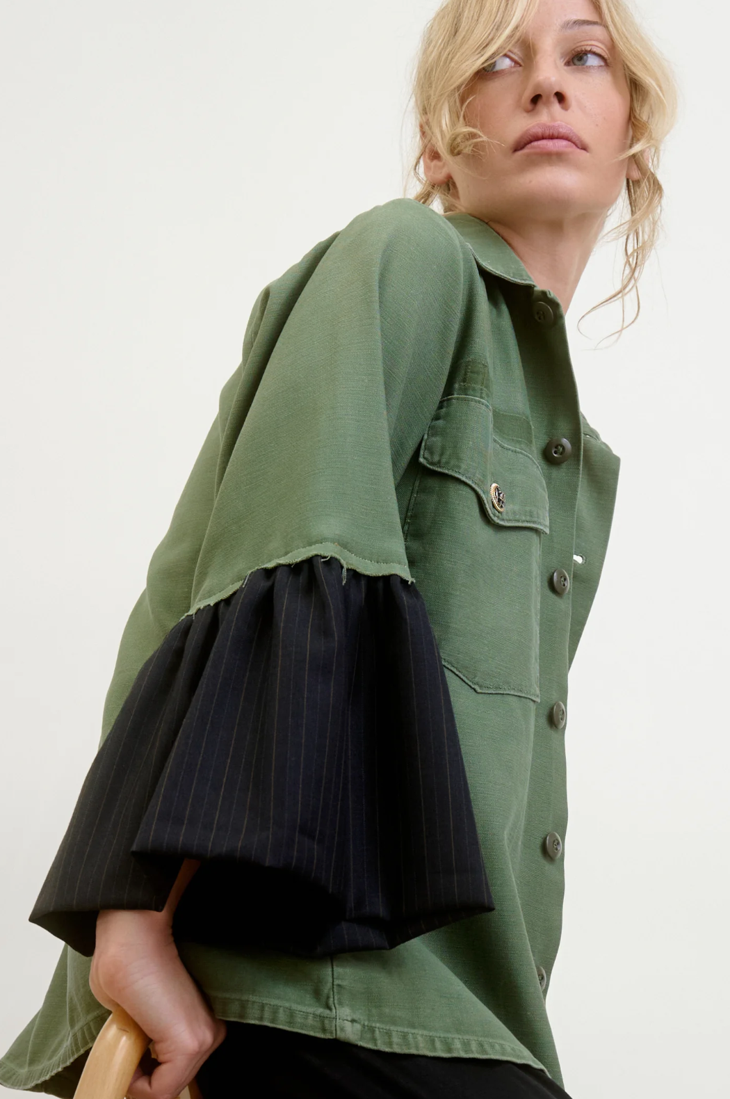 A person is wearing the Aquarius Cocktail Hooper Suiting Cuff Army Jacket, featuring black pinstriped bell sleeves. With loosely styled blonde hair conveying casual elegance, they stand against a white background, looking slightly upward.