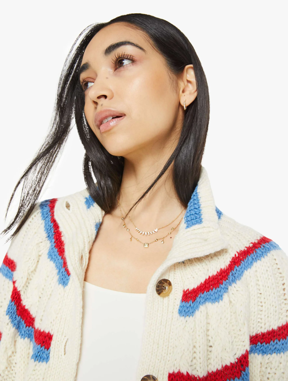 A woman with long dark hair wears The Mock Neck Button Cardigan by Mother, a slouchy fit in cream with red and blue stripes over a white top. Layered gold necklaces adorn her as she gazes upwards against a plain white background, adding elegance to her look.