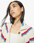 A woman with long dark hair wears The Mock Neck Button Cardigan by Mother, a slouchy fit in cream with red and blue stripes over a white top. Layered gold necklaces adorn her as she gazes upwards against a plain white background, adding elegance to her look.